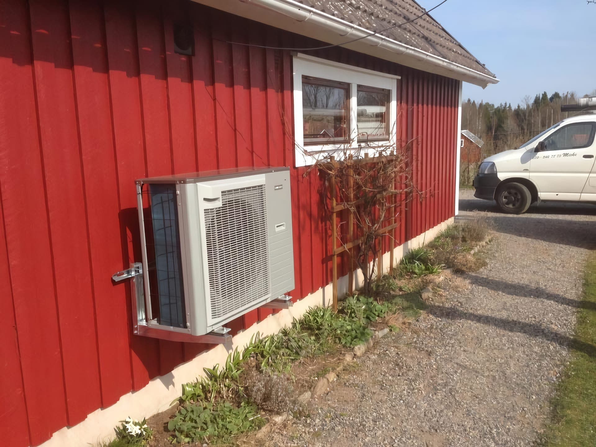 Grå luft/vatten-värmepump på en faluröd husvägg i vårsol, med Sandbergs Rörs servicebil i bakgrunden.