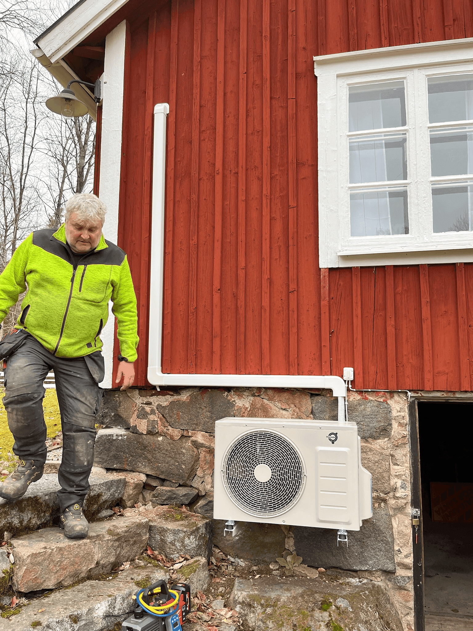 Montör intill en nyinstallerad luft/vatten-värmepump vid ett rött hus.